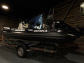 A black BRIG inflatable boat on a trailer, photographed inside a dark showroom or garage with wood-paneled walls.