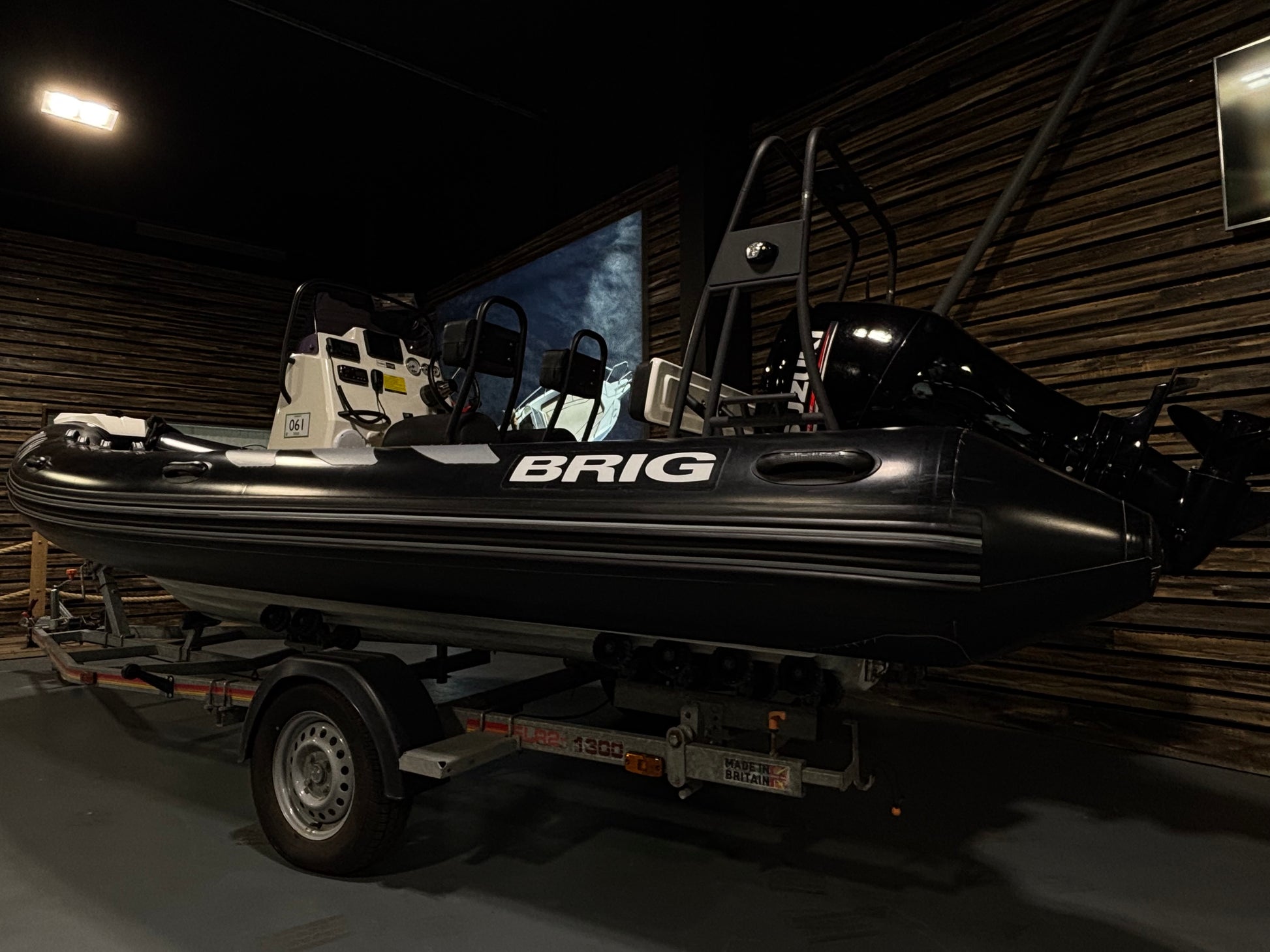 A black BRIG inflatable boat on a trailer, photographed inside a dark showroom or garage with wood-paneled walls.