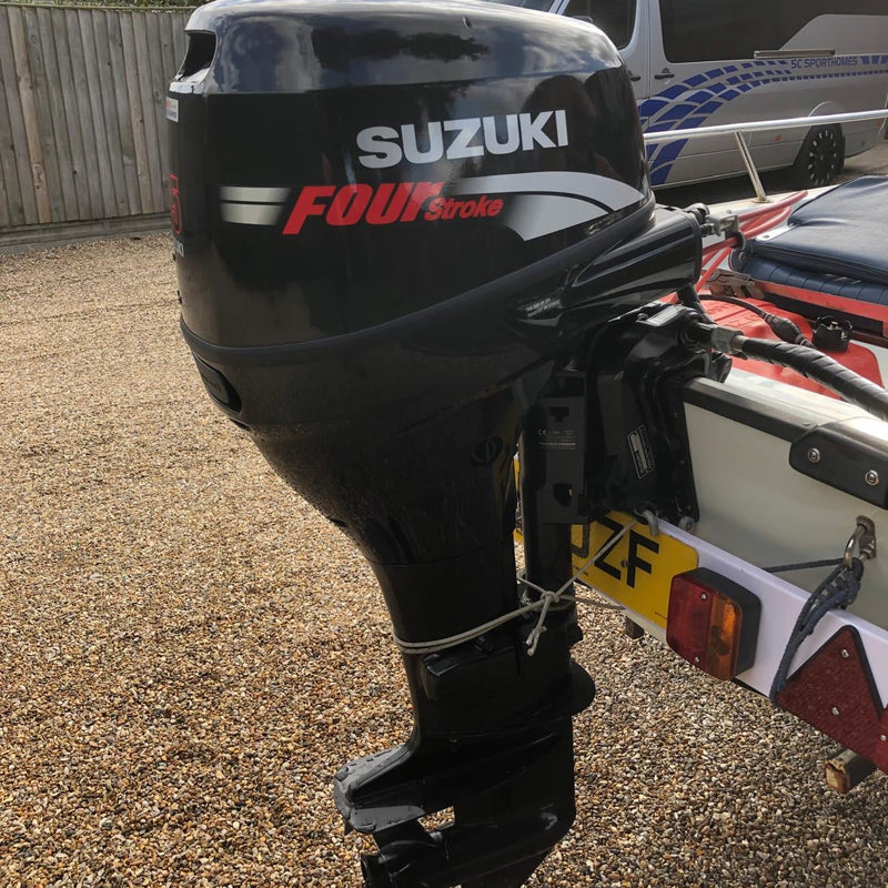 A black Suzuki FourStroke outboard motor mounted on a boat, photographed outdoors on a gravel surface with a white van in the background.