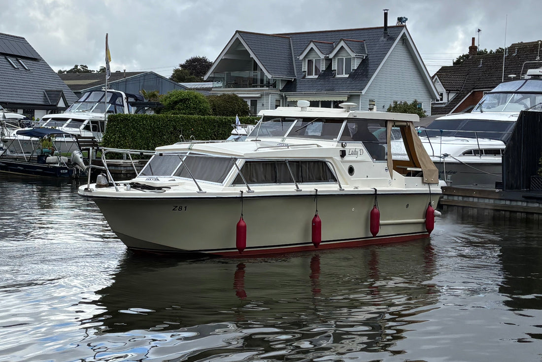 A cream-colored motor yacht with red fenders and a dark wood cabin frame, docked in a marina with houses in the background.